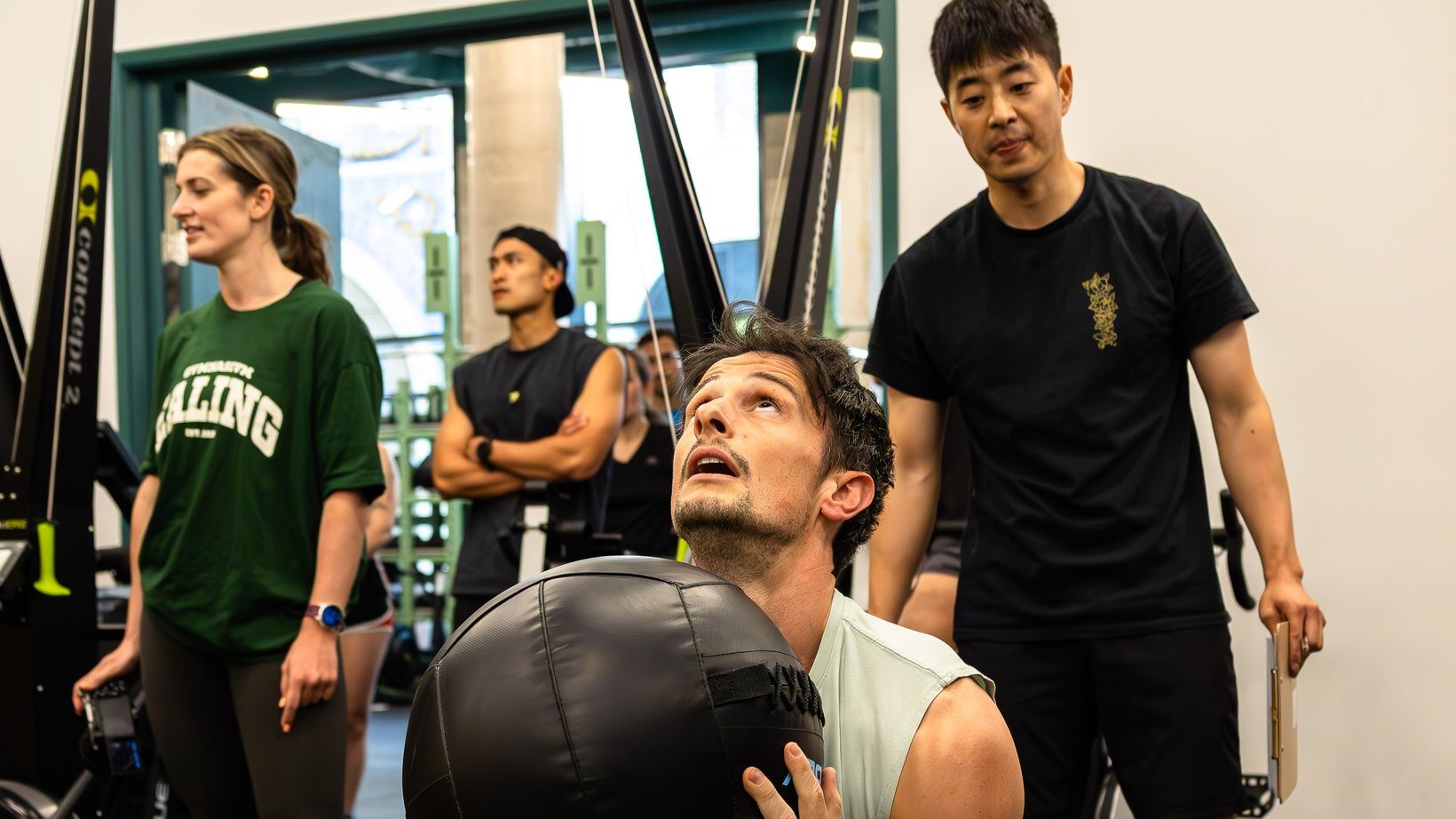 Member driving up out of a squat with a medicine ball, coach watching on